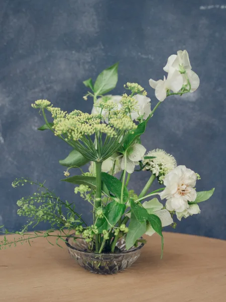 Wedding centerpiece, a low floral arrangement in a glass bowl, set on a wood tabletop against a soft blue wall