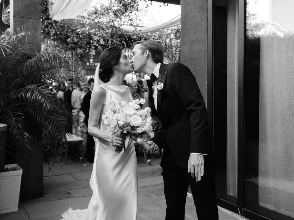 Wedding kiss portrait during a ceremony recessional kiss, bride clutching a bridal bouquet as guests cheer beneath a draped canopy on a stone patio.