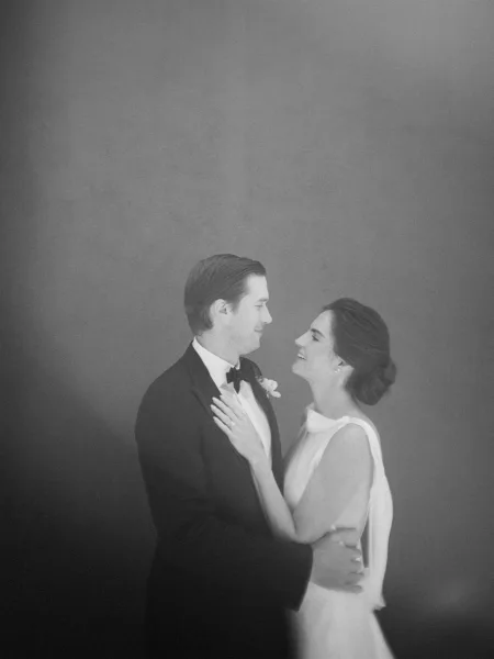 Couple portrait as bride and groom embrace, he holds her close, her bridal updo framed softly against a minimalist studio backdrop