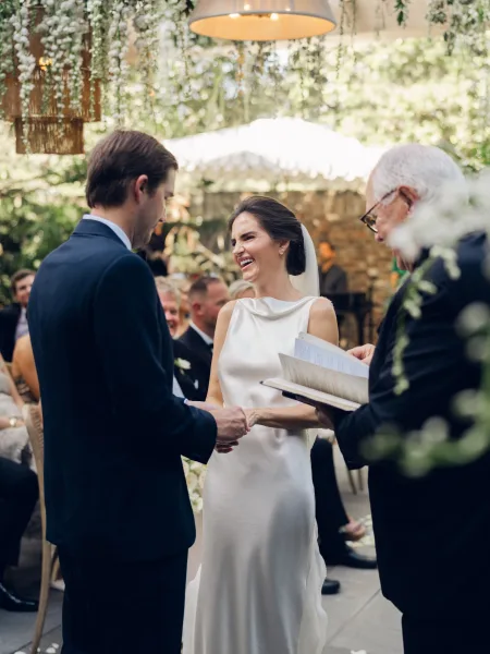 Ceremony moment during wedding vows as the bride and groom hold hands, with the officiant reading from a book, in a garden setting.