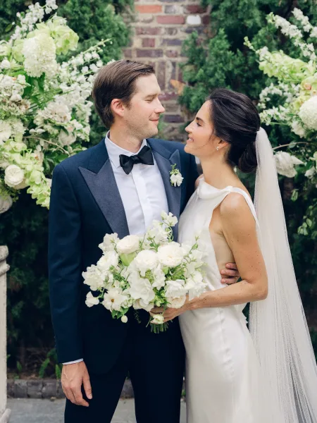 Couple portrait of bride and groom smiling, with the bride holding a white bouquet against a brick wall and garden greenery.