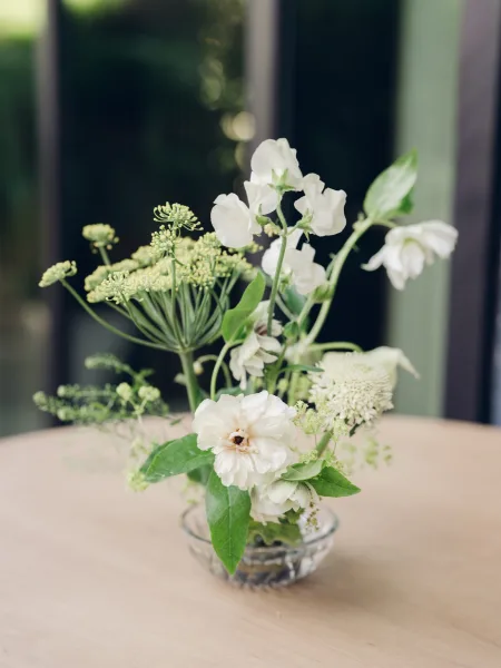 Wedding centerpiece, a white flower centerpiece on a light wood table by a window with garden greenery outside as a natural backdrop.