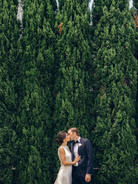 Wedding kiss portrait as a bride and groom kiss, her satin bridal gown catching light before an evergreen hedge and tall trees.