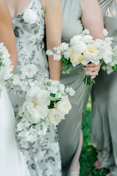 Bridesmaid bouquets of white roses with ranunculus and sweet peas, tied with greenery, held by bridesmaids in sage dresses on a lawn
