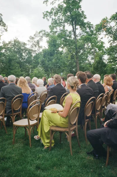Ceremony guests fill outdoor ceremony seating, perched on bentwood chairs beneath trees on a grass lawn.