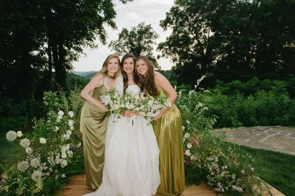 Bride and bridesmaids in an outdoor bridal party portrait, smiling on a garden hillside with trees and sky, the bride holding a lush bridal bouquet.