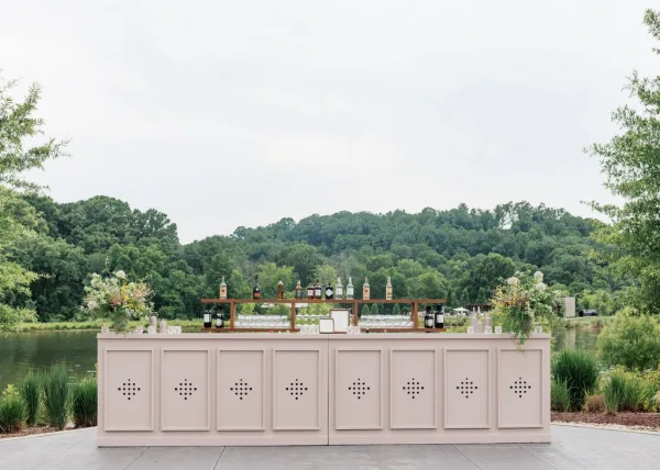 Wedding bar setup as an outdoor bar station with soft florals, arranged on a concrete patio beside a calm lake with trees and rolling hills.