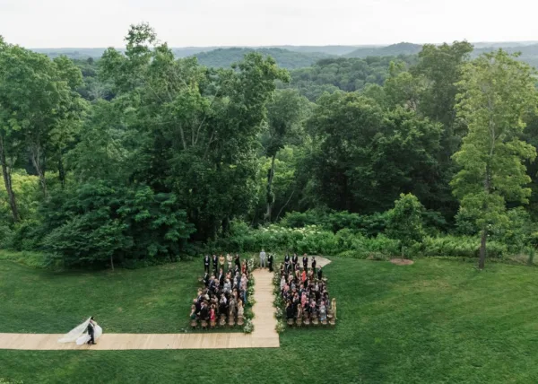 Ceremony moment as the couple is walking down the aisle, her bridal veil trailing, on a lawn framed by trees and distant forest hills.