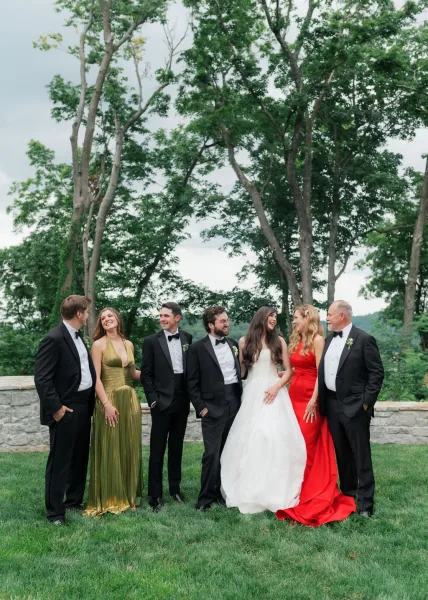 Wedding party portrait laughing together in an outdoor group wedding photo by garden trees and a stone wall, with crisp white boutonnières.