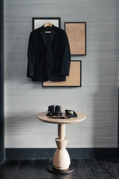 Groom attire featuring a black tuxedo and bow tie against a textured wall with framed artwork and wood floor