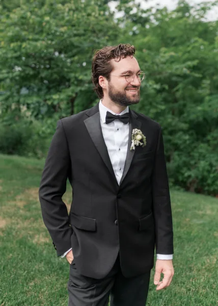 Groom portrait of a black tux groom smiling in a tailored tux with a white boutonniere, set against garden trees.