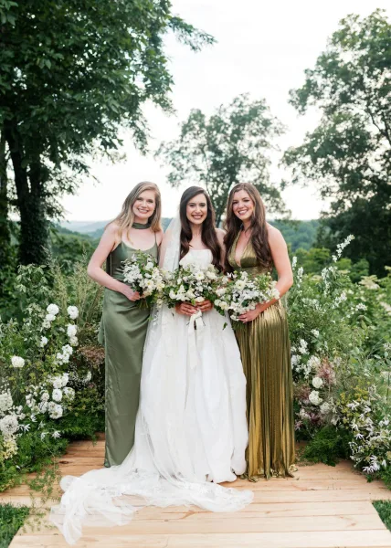 Bride with bridesmaids in green bridesmaid dresses stand on a wooden platform, holding bouquets amid garden greenery and trees.