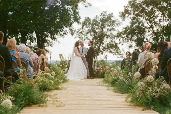 Ceremony moment with the couple exchanging vows on a wooden aisle, surrounded by trees under open sky with distant hills framing the scene.