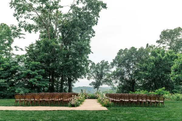 Ceremony setup with outdoor ceremony seating in symmetrical rows of bentwood chairs along a simple aisle on a tree-lined lawn with distant hills.
