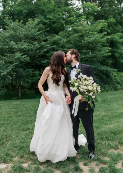 Wedding kiss portrait of newlyweds sharing a tender moment in a garden, the bride cradling a white bridal bouquet.