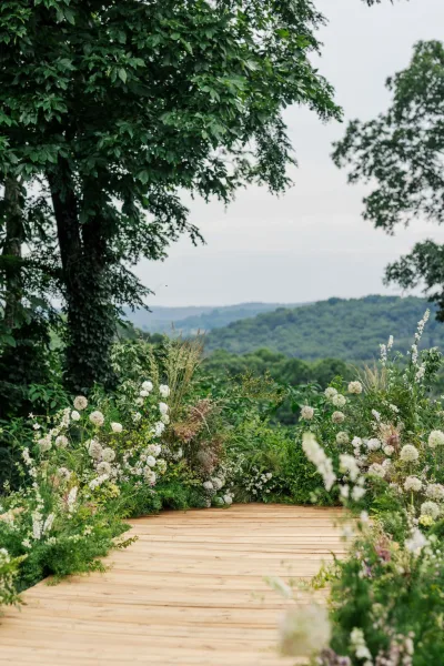 Ceremony aisle design with outdoor ceremony decor on a wooden platform, set before rolling hills and trees beneath an open sky.