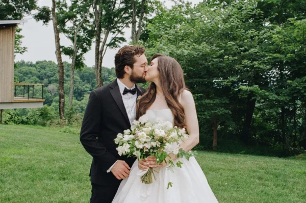 Wedding kiss portrait as the bride and groom kiss outdoors, she holds a bridal bouquet beneath shady garden trees.