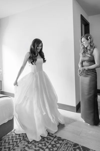 Bride getting ready during a wedding morning moment, smoothing a satin gown indoors near a doorway, by a white wall.