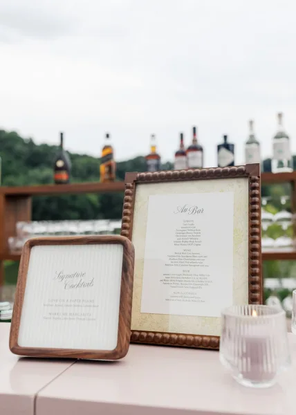Wedding bar signage with a signature cocktail sign in wood frames, styled on a wooden bar shelf with trees and a cloudy sky beyond.