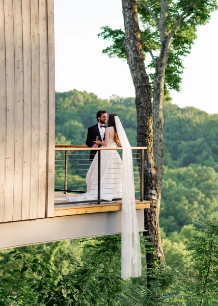 Couple portrait on a balcony, an outdoor wedding portrait as they raise champagne glasses, with forested hills and sky stretching beyond.