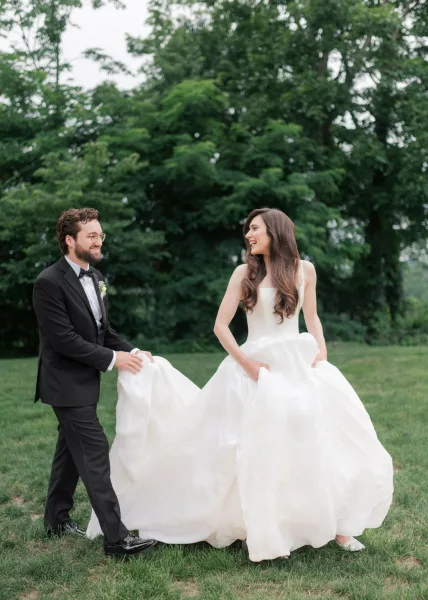 Couple portrait of the bride and groom walking across a garden lawn, her dress swaying as the groom in eyeglasses smiles warmly.