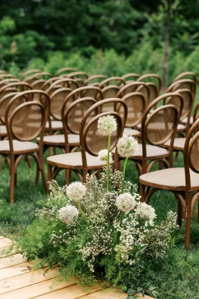 Ceremony aisle flowers shape an outdoor ceremony setup, featuring white allium on a garden lawn bordered by trees.