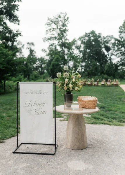 Wedding welcome sign serving as the ceremony entrance sign on a metal sign stand, set on a grass lawn beside garden chairs and trees.