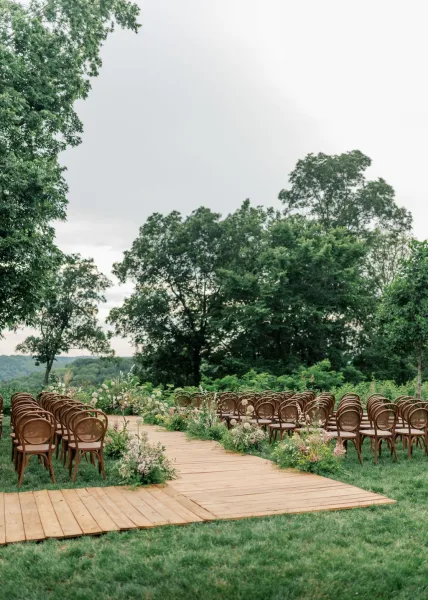 Ceremony setup for an outdoor garden ceremony with ground florals lining the aisle on a grass lawn under garden trees and a soft cloudy sky.