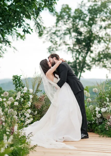 Ceremony kiss moment during the wedding first kiss as the couple embraces, her veil catching light, with distant hills and trees under open sky.