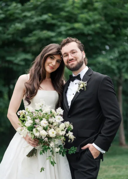 Couple portrait, an outdoor wedding portrait of a bride and groom smiling as she holds a white and green bridal bouquet amid garden trees.