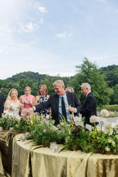 Escort card display on an outdoor escort card table adorned with string lights, set against blue sky, garden trees, and hills.