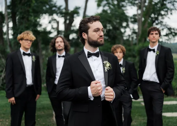 Groom portrait with a bow tie, standing on a garden path amid trees and a green lawn under a softly cloudy sky.