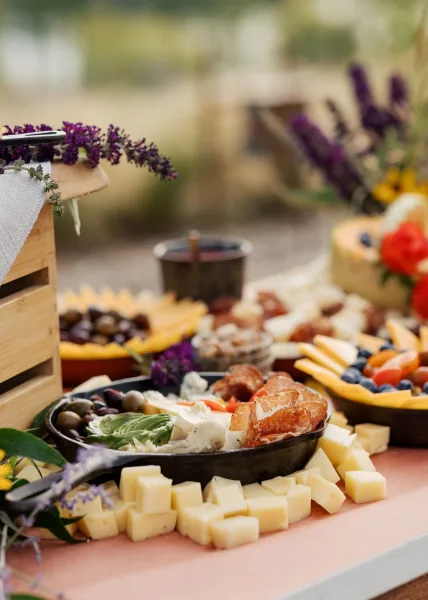 Charcuterie board styled for a wedding grazing table, featuring cheese cubes on an outdoor table amid garden greenery and blurred florals.