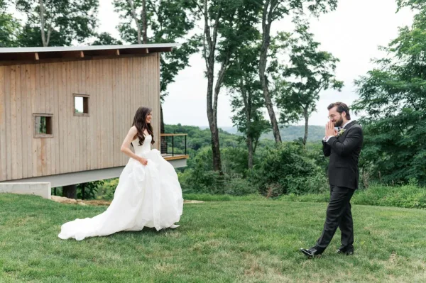 Wedding first look with groom reaction as the bride approaches in a flowing bridal gown on a grass lawn by a wood building and distant hills.