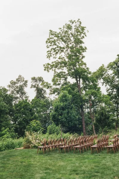 Ceremony setup for outdoor ceremony seating with rows of wooden chairs on a grass lawn framed by garden trees under a clear sky.
