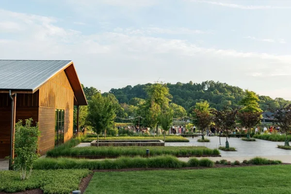 Outdoor wedding venue with string lights above a courtyard beside a modern barn, backed by trees and rolling hills.
