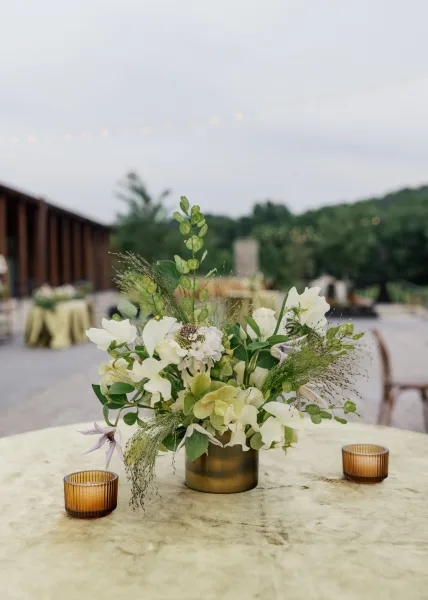 Wedding centerpiece, a cocktail table centerpiece in a gold vase, set on an outdoor patio beneath string lights and garden trees.