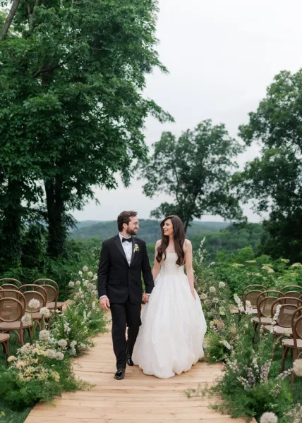 Couple portrait of newlyweds, couple holding hands and gazing at each other on a wooden aisle, framed by garden trees with distant hills beyond.