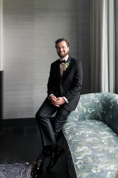 Groom portrait of a black tuxedo groom seated on a patterned sofa, framed by window curtains and a textured wall in an indoor setting.