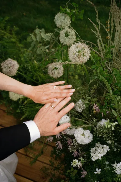 Wedding rings on bride and groom hands, a diamond engagement ring shown as they rest gently amid garden greenery.