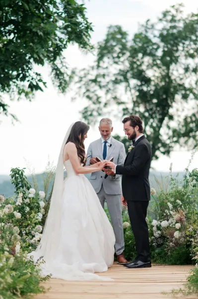 Wedding ring exchange during an outdoor ceremony moment as the bride slides his band, an officiant book open on a wooden platform.