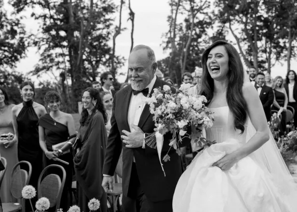 Ceremony processional with the bride walking down the aisle, carrying a bridal bouquet, as guests look on beneath trees.