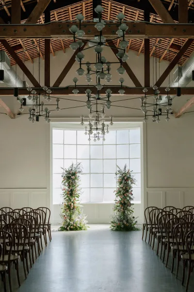 Ceremony setup with floral pillars framing a modern ceremony aisle, wooden chairs and chandeliers in an airy loft with grid windows