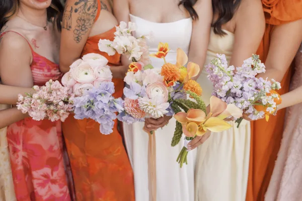 Bridesmaid bouquets in colorful bridesmaid bouquets palette, hand-tied with ranunculus and ribbons, held by bridal party torsos on neutral backdrop