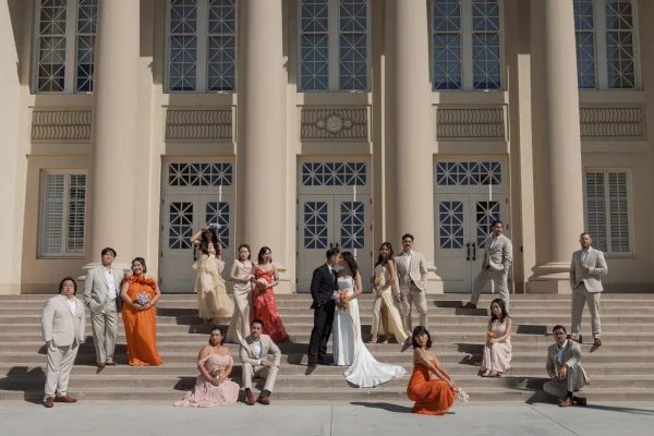 Wedding party portrait with bride holding bouquet and groom in black tux, bridesmaids and groomsmen posed on stone steps with columns backdrop