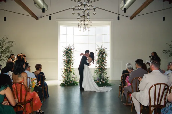 Ceremony kiss as the bride in a strapless wedding dress and groom in black suit kiss beneath floral pillars, guests watching in modern venue