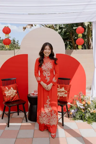 Bridal portrait of a red ao dai bride in an embroidered dress seated by red lanterns and tea set on a patio with brick wall greenery