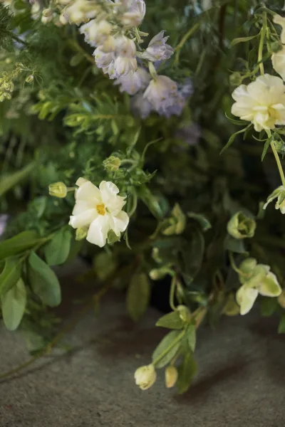 Wedding florals with white blooms, pale purple buds, and lush greenery arranged on a textured stone surface with soft foliage accents