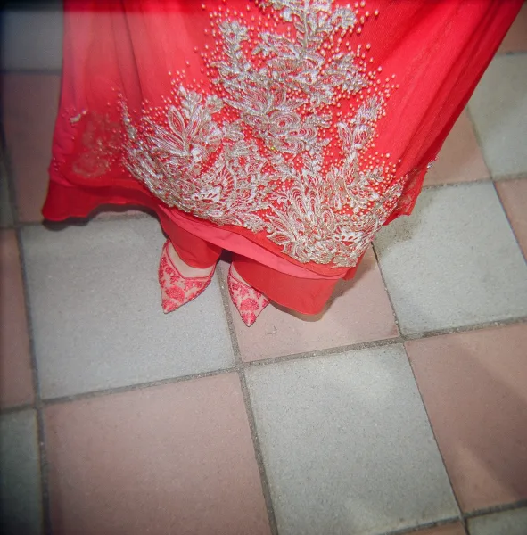 Wedding shoes, red wedding heels with pointed toes and ankle straps, beside a beaded gold-embroidered dress hem on stone tile floor