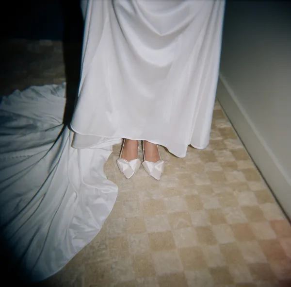 Bridal shoes with bows peeking from under a satin wedding dress hem and train, pointed white heels on a tile hallway floor